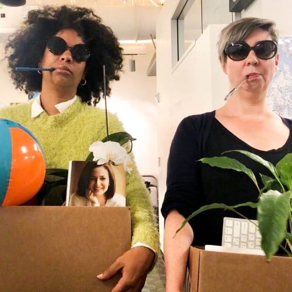 Two women wearing sunglasses walk side-by-side, each holding bankers' boxes.