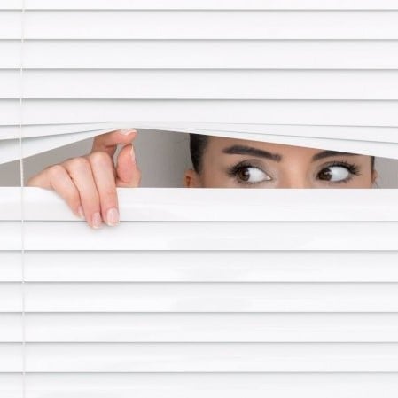 A par of eyes peeks out from between venetian blinds propped open by a hand.