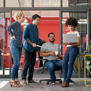 A group of employees wearing jeans cluster around a desk in an office