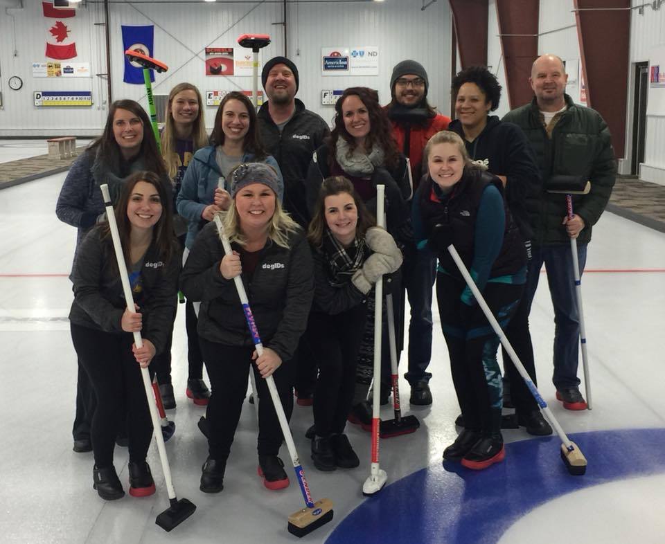 dogIDs team members pose with curling brooms at a curling rink.