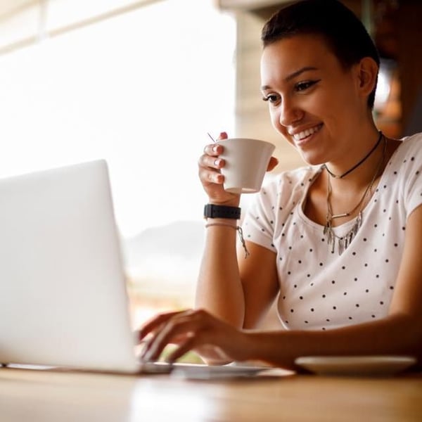 A woman smiling and holding a cup while looking at a laptop.