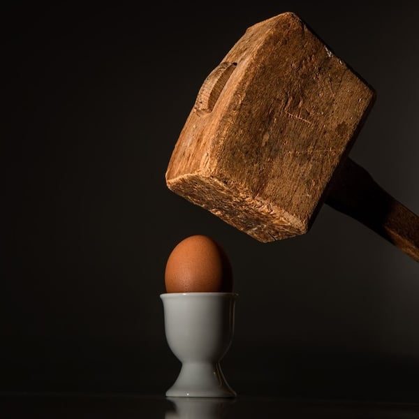 A mallet poised above a brown egg in an egg cup.