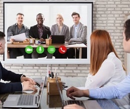 Coworkers place a video call from office to office, with workers at a conference table looking at a screen featuring workers on a wall.