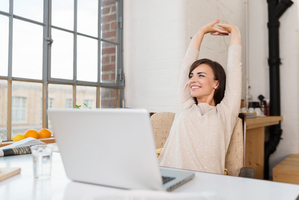 Happy relaxed young woman sitting at a desk with a laptop in front of her stretching her arms above her head and looking out of the window with a smile